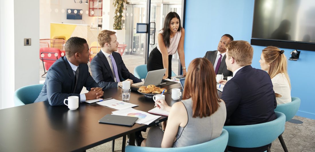 Female manager stands addressing colleagues in meeting room